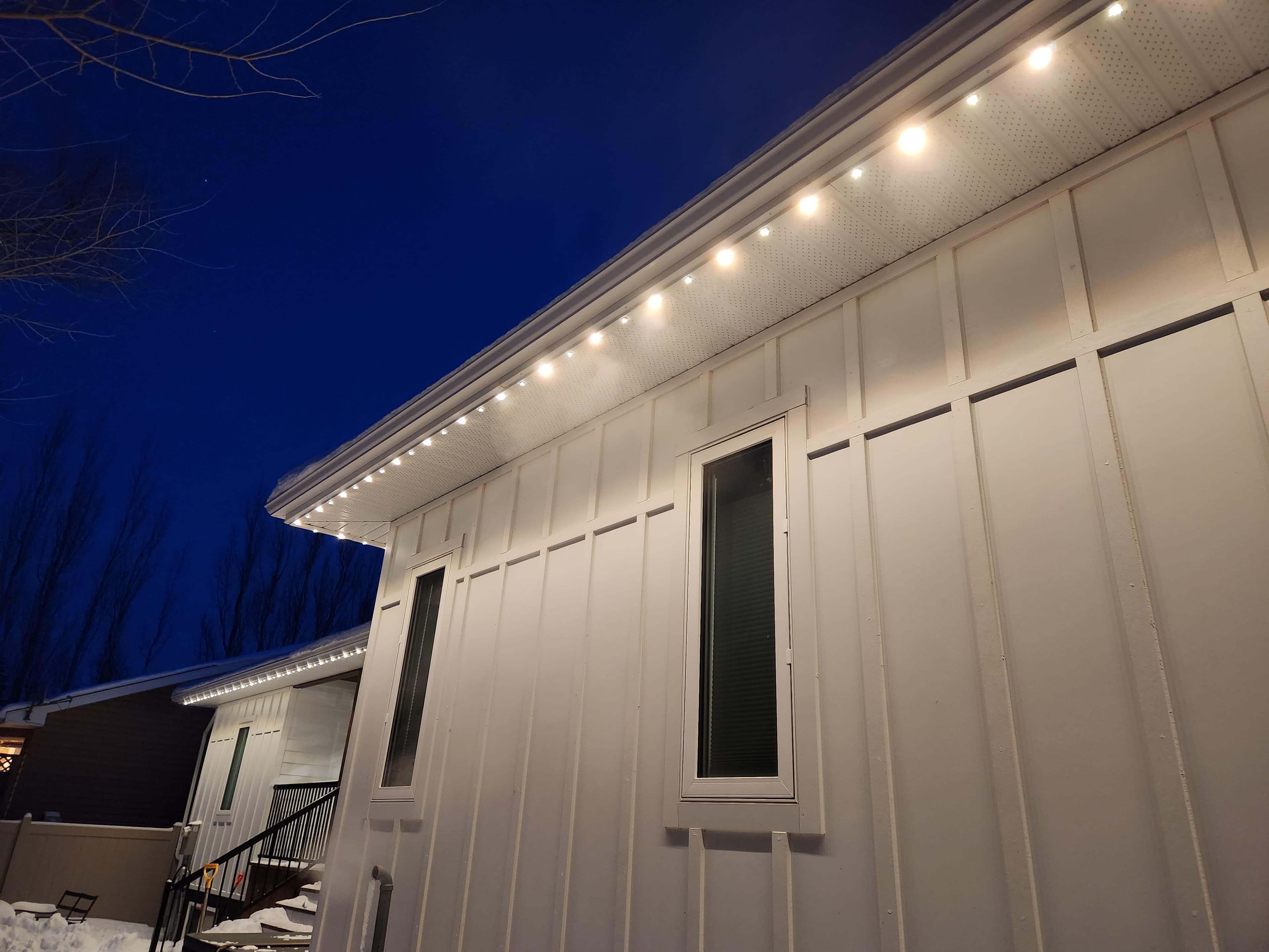 Close-up of permanent LED nodes installed under the soffit of a white board-and-batten home in winter